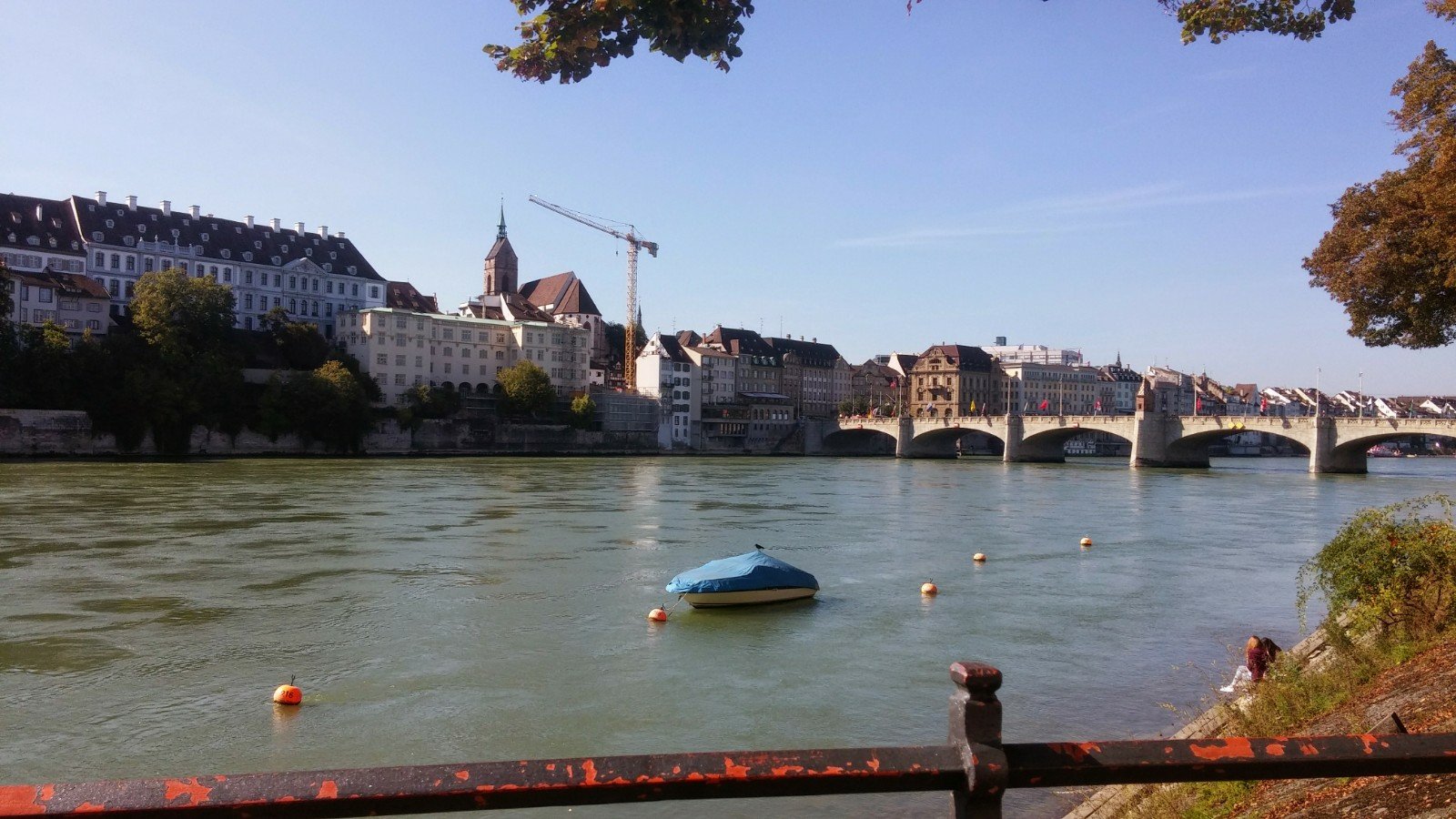 Rijnfietsroute EV15 vanaf Basel. River with a small covered boat and buoys, buildings along the riverbank, and a stone bridge in the background under clear sky