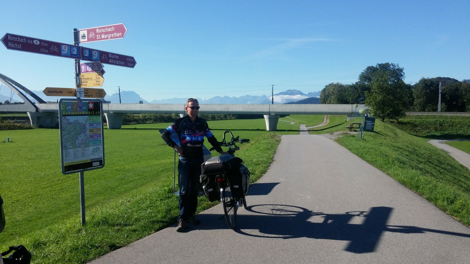 A cyclist in a biking outfit stands beside a bicycle on a path surrounded by green grass. Nearby, there are direction signs indicating various routes and distances, with a backdrop of mountains and a clear blue sky.