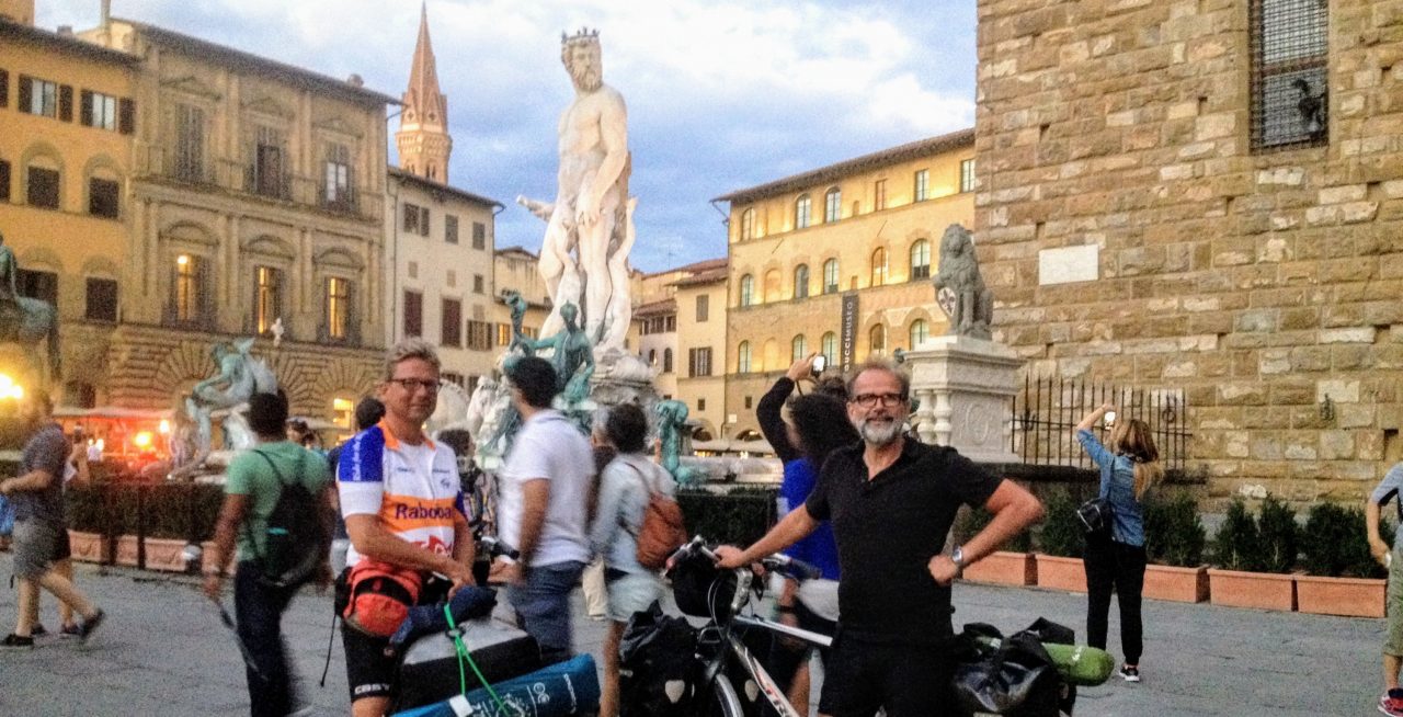 Two cyclists posing in front of a famous statue in a busy plaza, with people walking around and historic buildings in the background.