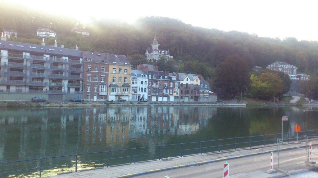 A scenic river view featuring a mix of modern and traditional buildings along the water's edge, with trees and a hillside in the background.