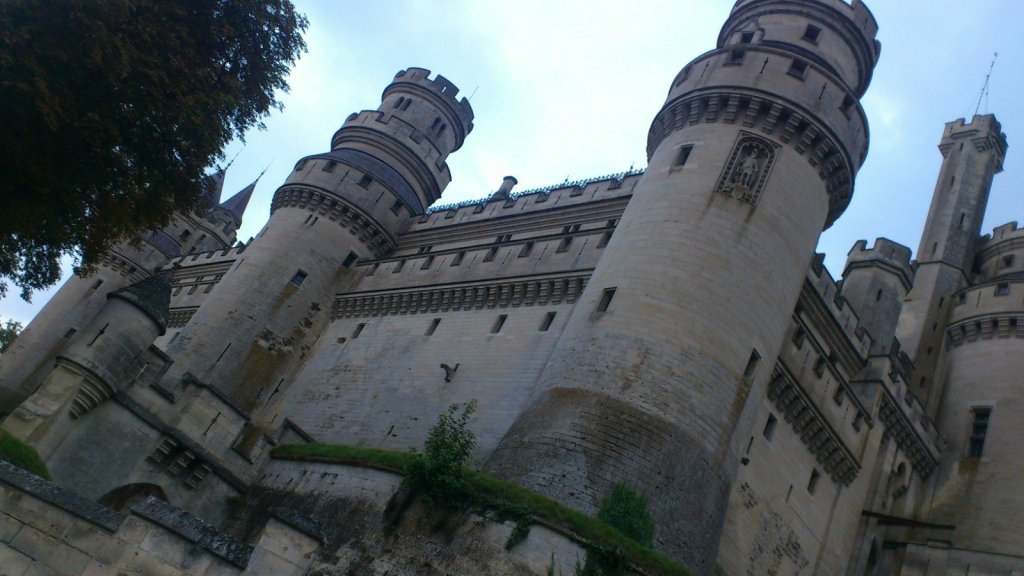 Angled view of a historic castle with towers and intricate stonework, surrounded by greenery.