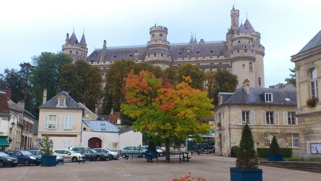 A picturesque view of a castle with turrets set against a cloudy sky, surrounded by trees and autumn foliage, in a town square featuring parked cars and buildings.