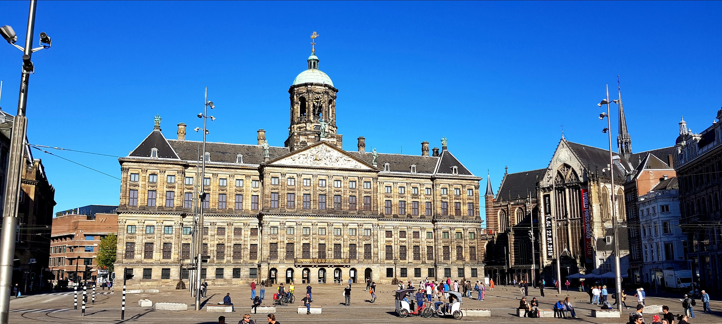 Historic building in Amsterdam with a large dome and clock tower, surrounded by a lively square with people and clear blue skies.