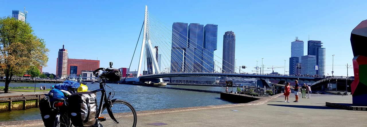 soorten fietsvakanties Bicycle with packed panniers parked near a river with a cable-stayed bridge and modern buildings in the background