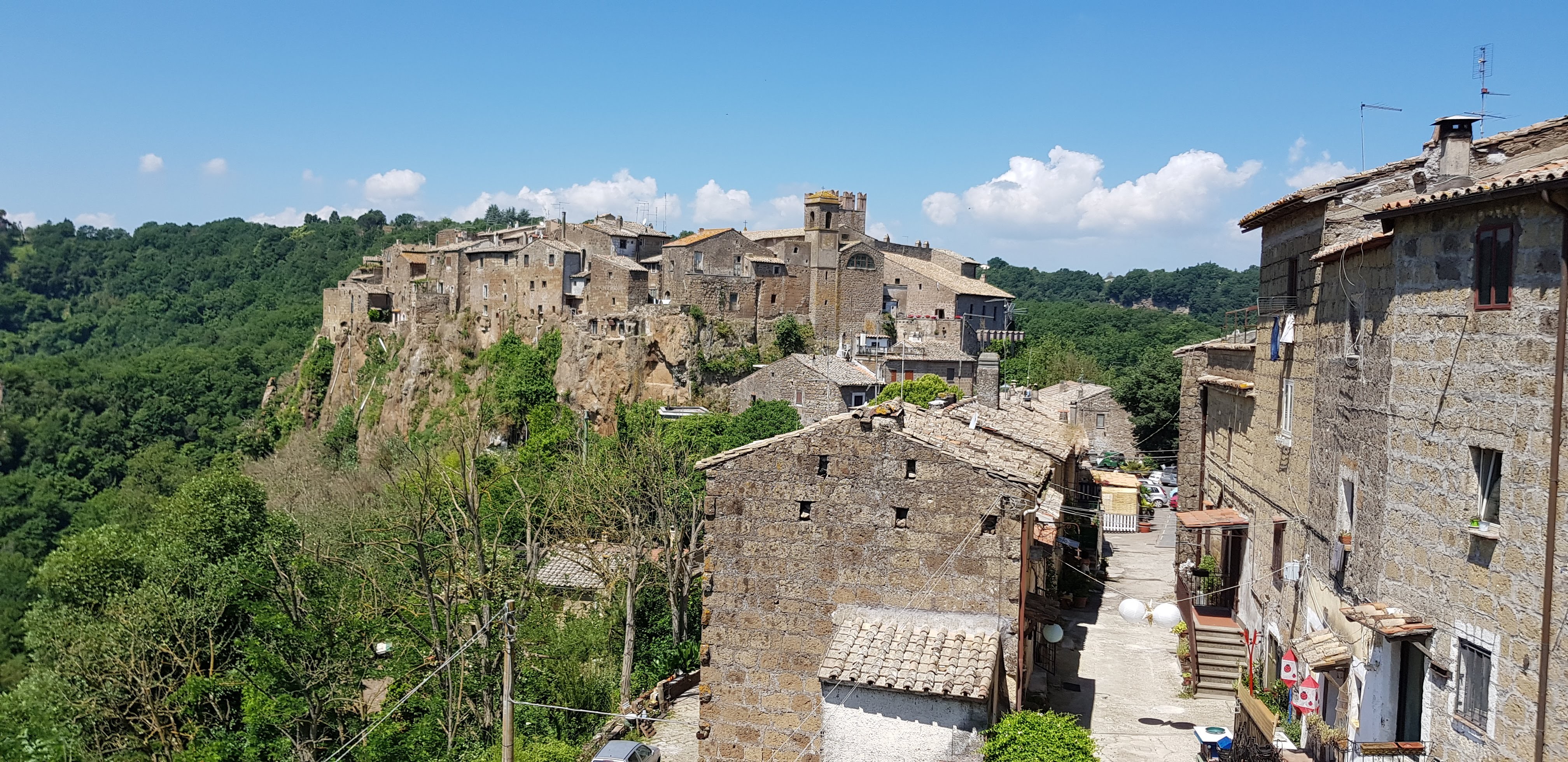A panoramic view of an ancient village perched on a cliff, surrounded by lush greenery and under a clear blue sky.