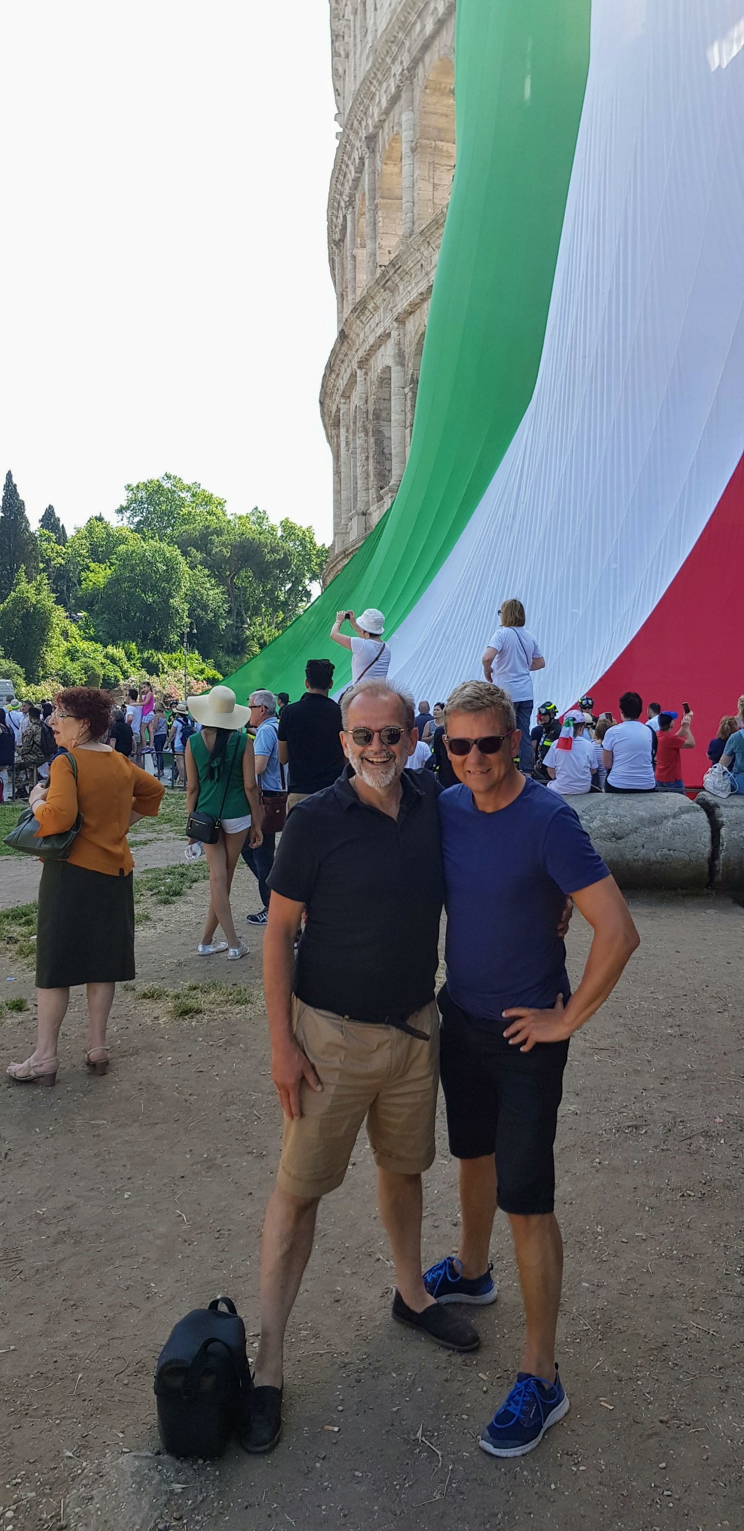 Two men pose in front of a large Italian flag draped near the Colosseum in Rome, with a crowd of people in attendance.