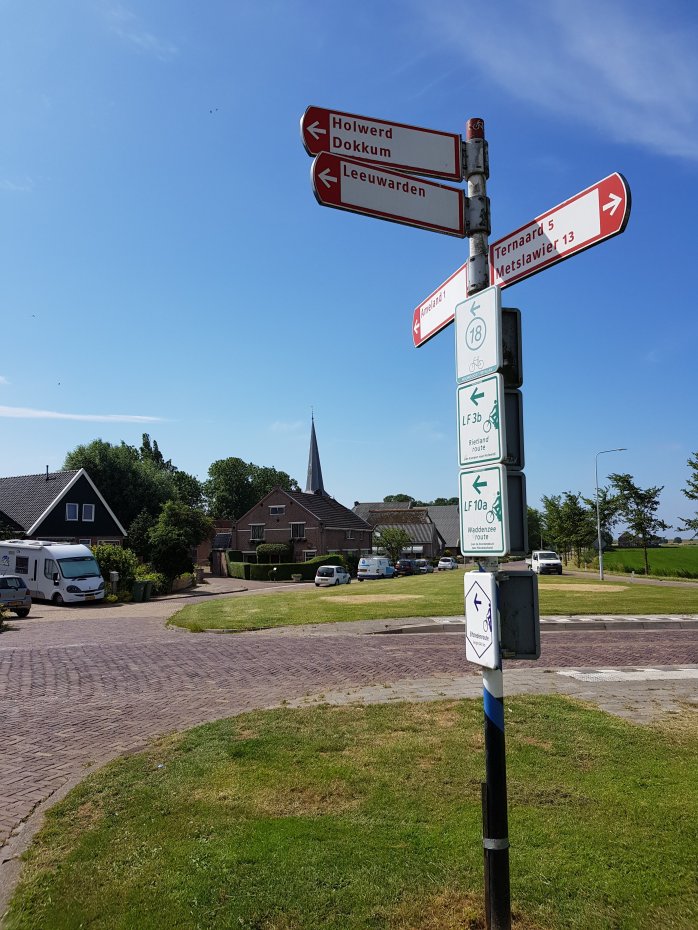 A directional signpost indicating routes to Holwerd, Dokkum, and Leeuwarden, set against a blue sky with a village in the background.