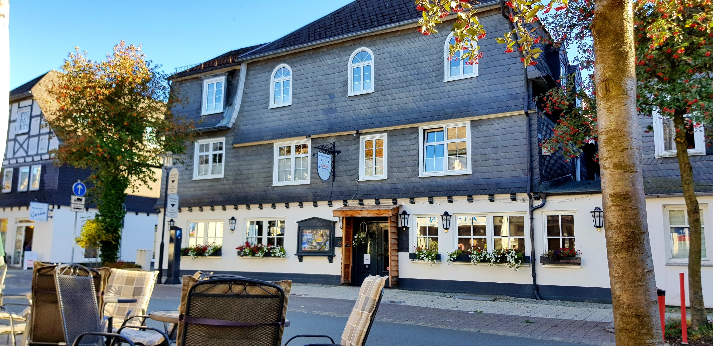 A charming two-story building with a gray shingle exterior, featuring multiple windows and flower boxes. There's a wooden entrance and decorative lampposts. In the foreground, there are outdoor seating arrangements with metal chairs.