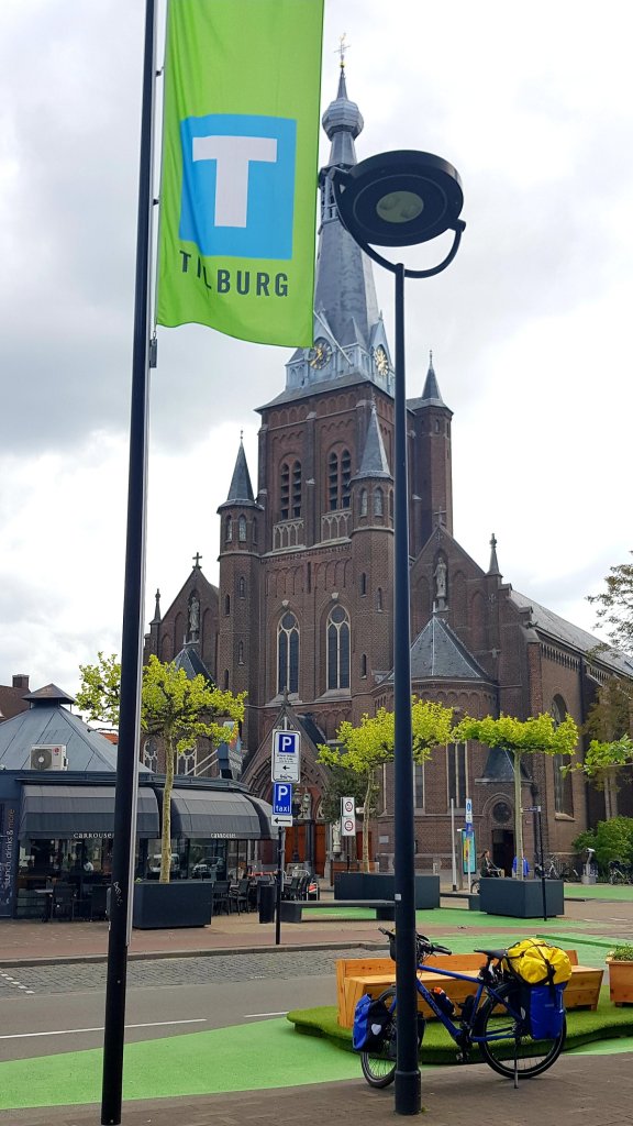 A view of a tall brick church with spires and a clock tower, accompanied by a green flag displaying the word 'TILBURG'. A parked bicycle with yellow bags is in the foreground, along with outdoor seating and trees in a city square.