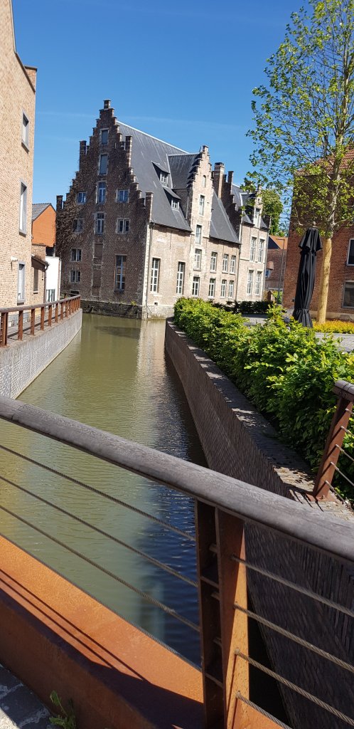 A scenic view of a narrow canal surrounded by historic brick buildings and lush greenery under a clear blue sky.