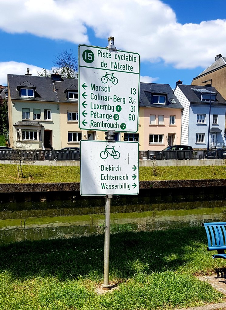 Signpost for the Alzette cycling path with directions to various towns including Mersch, Colmar-Berg, and Diekirch, surrounded by green grass and houses under a blue sky.
