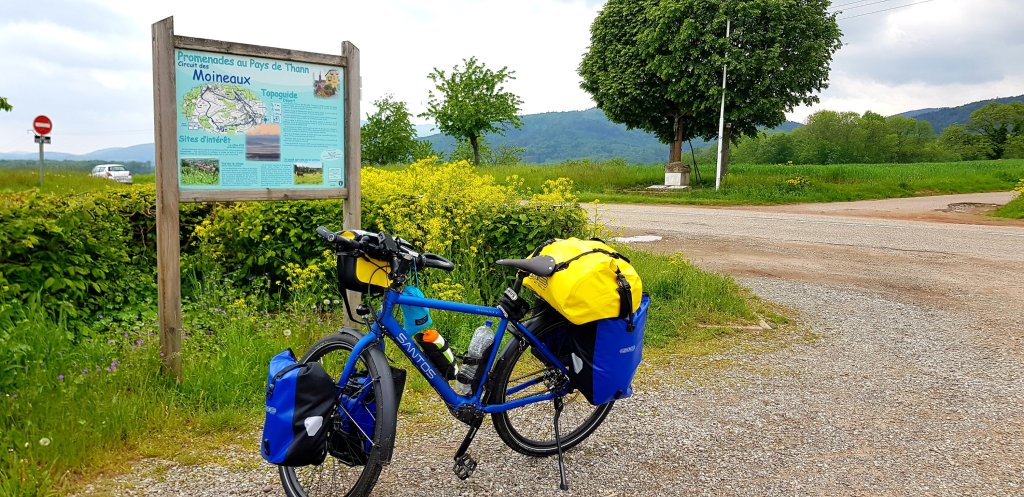 A blue touring bicycle with yellow and blue panniers parked near a sign for hiking routes in the Pays de Thann region, surrounded by greenery and flowers.