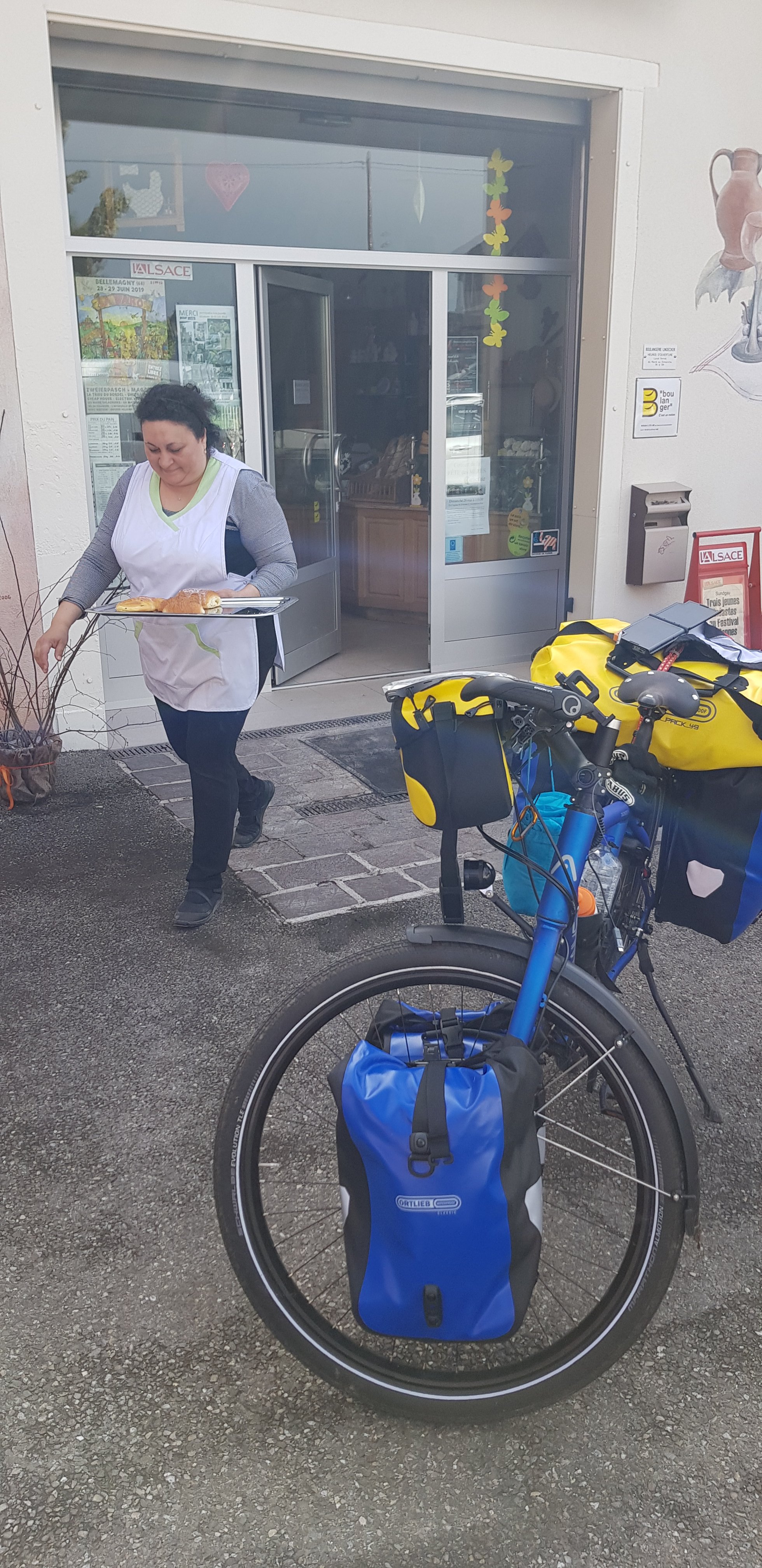 Een vrouw in een schort loopt naar buiten met een dienblad in de hand, terwijl een fiets met bagage naast haar staat.