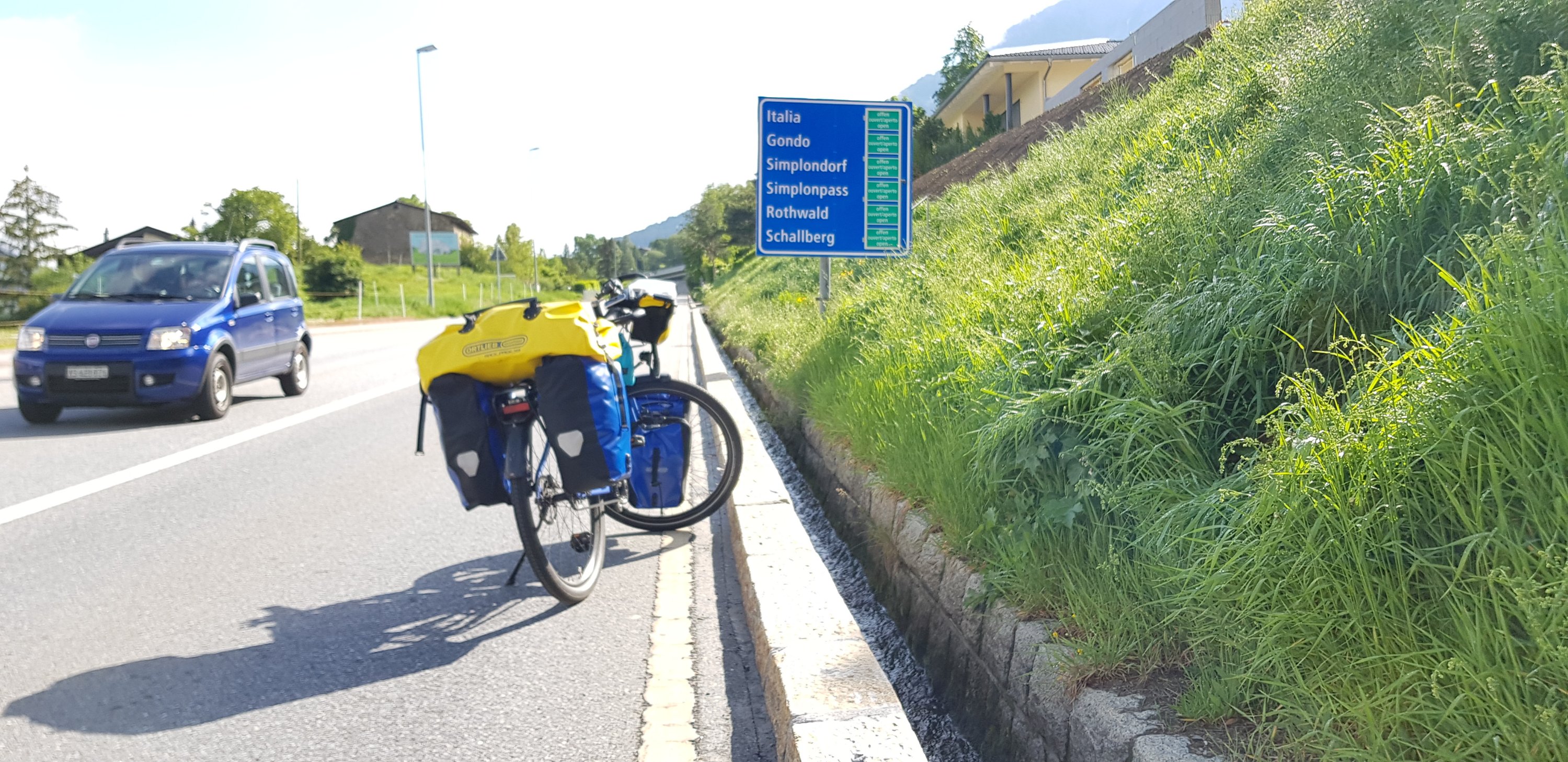 A bicycle with yellow and blue panniers parked near a roadside sign indicating directions to various locations including Italia, Gondo, and Simplonpass.