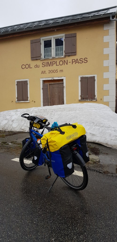 A blue and yellow bicycle with panniers parked in front of a building featuring a sign that reads 'COL du SIMPLON-PASS Alt. 2005 m', with snow in the background.