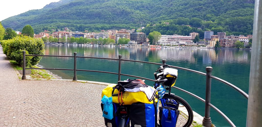 A bicycle with panniers is parked along a scenic lakeside path, overlooking a calm lake surrounded by lush green mountains and quaint buildings reflecting in the water.
