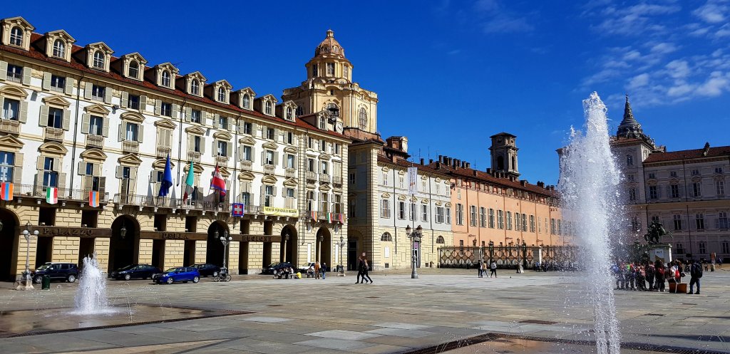 Wide view of a historic square in Turin, Italy, featuring ornate buildings, colorful flags, and a fountain, under a clear blue sky.