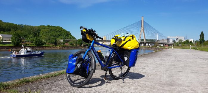 A blue touring bicycle with yellow panniers parked by a river, with a bridge and greenery in the background.