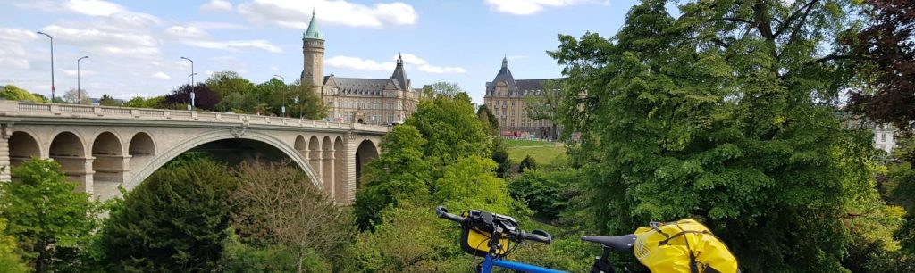 A view of a lush green landscape featuring a stone bridge, a blue bicycle with yellow bags in the foreground, and a historic building in the background under a partly cloudy sky.
