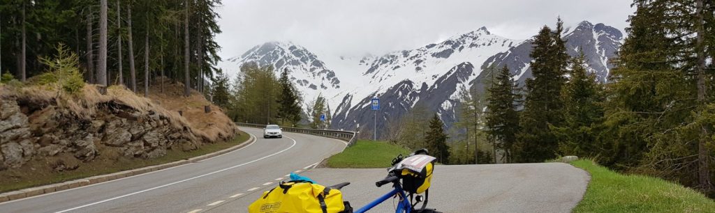 A bicycle with yellow panniers parked beside a winding road, with snow-capped mountains and dense green forest in the background under a cloudy sky.