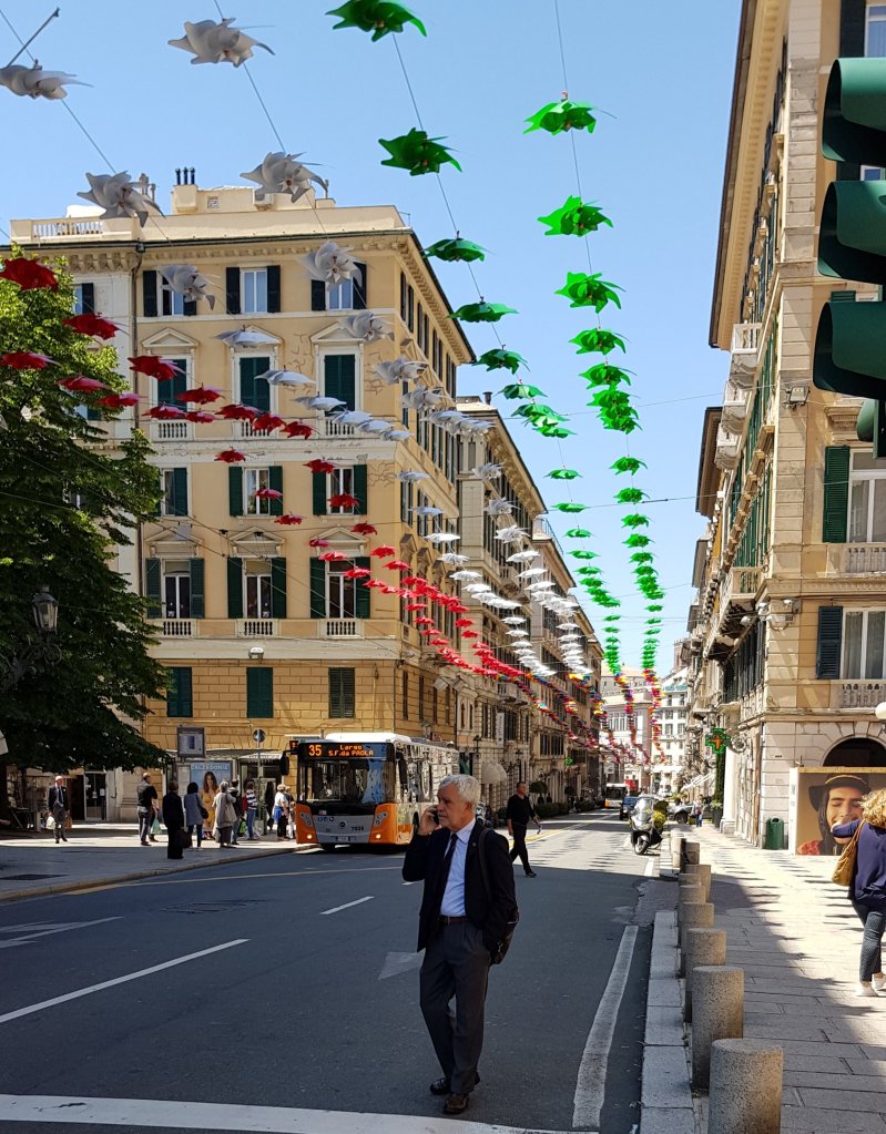 A busy street scene featuring a man on the phone, with colorful decorations hanging above, including red, green, and white umbrellas and flowers, and a bus in the background.