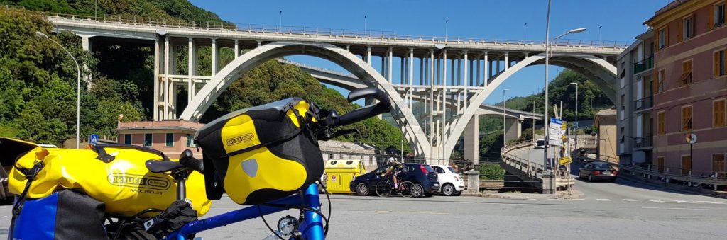 A blue bicycle with yellow Ortlieb bags parked in the foreground, featuring a large bridge and green hillside in the background.