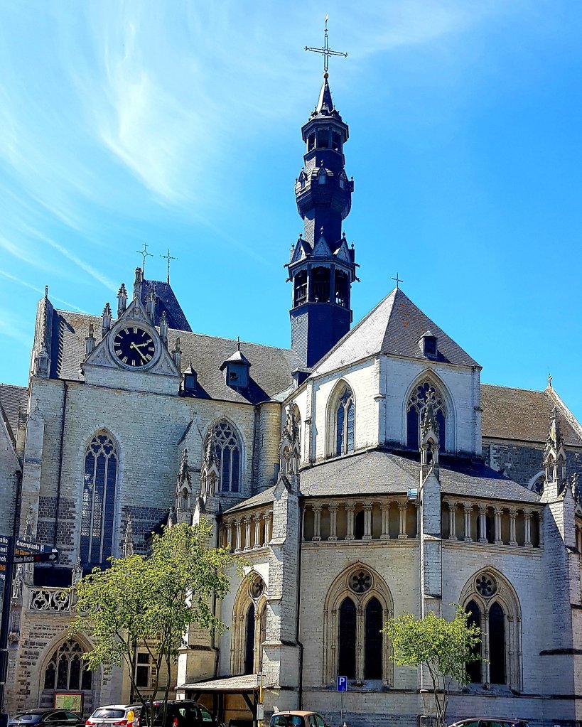 Exterior view of a historic church with a tall, ornate tower, intricate stonework, large windows, and a clock on the facade, set against a clear blue sky.