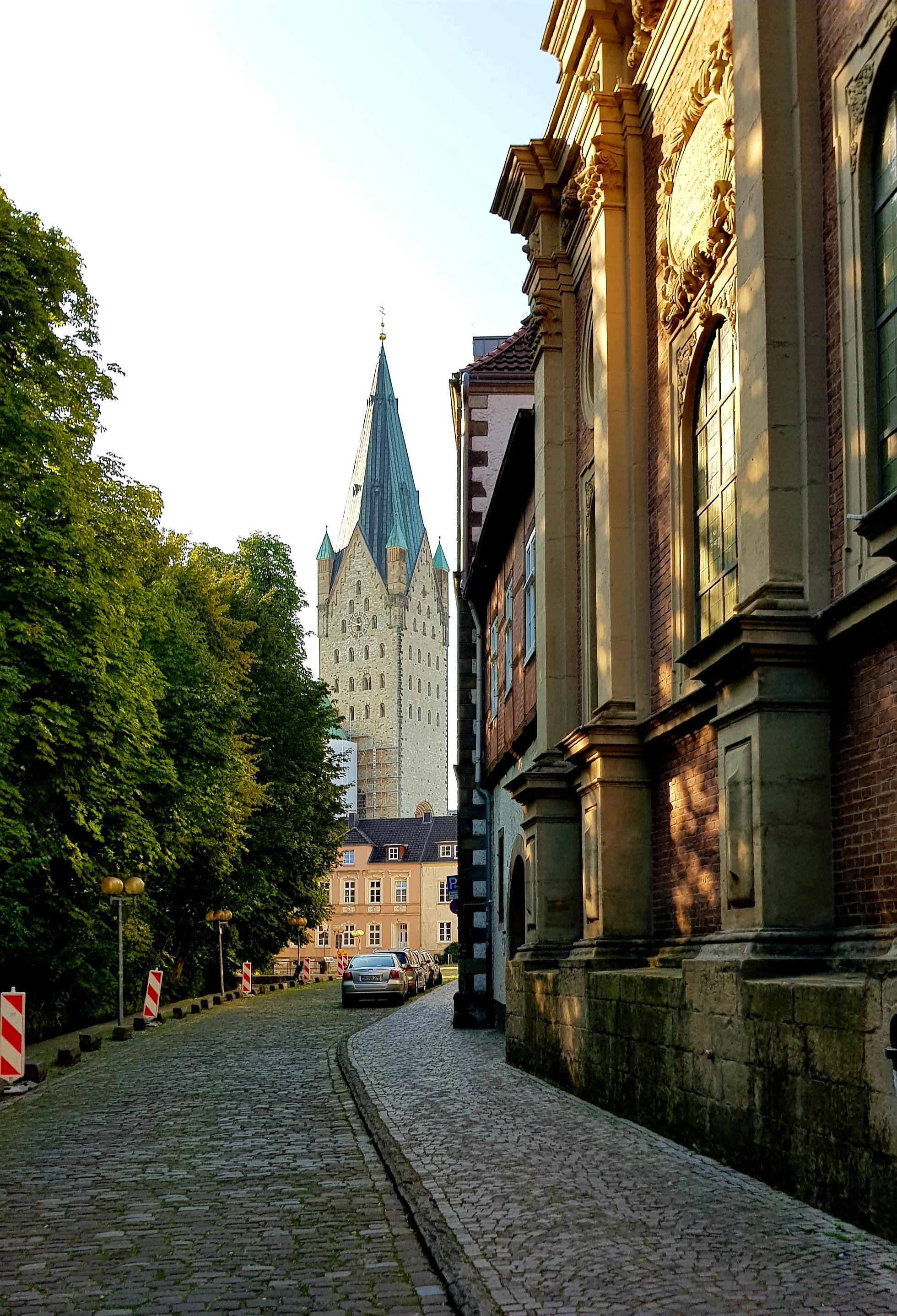 A cobblestone street lined with trees and historic buildings, featuring a prominent tower with a green, triangular roof in the background.