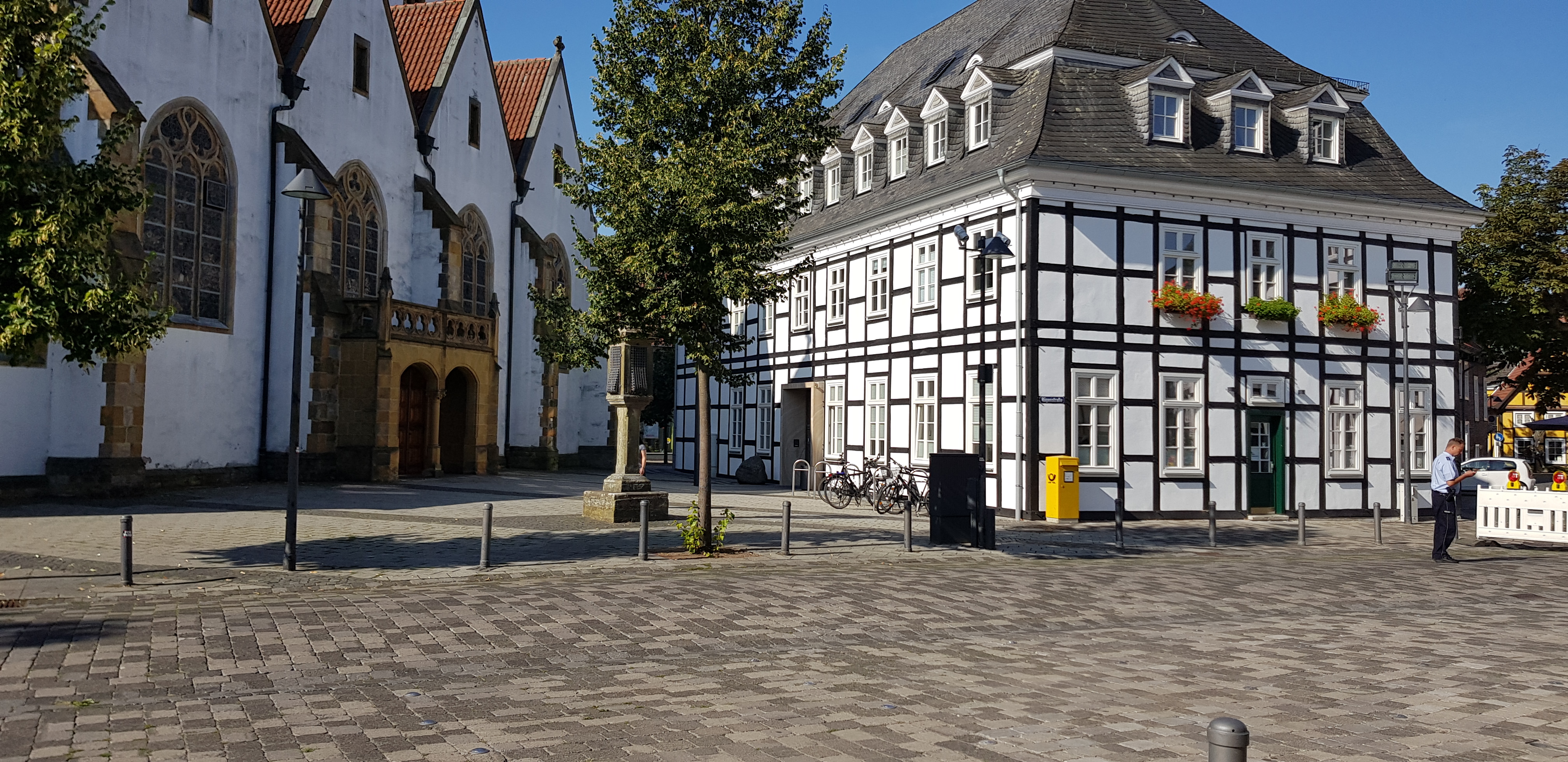 A quaint town square featuring a historic white building with tall windows and a distinctive roof, alongside another building with traditional half-timbered architecture. There are trees, lampposts, and bicycles parked near the square.