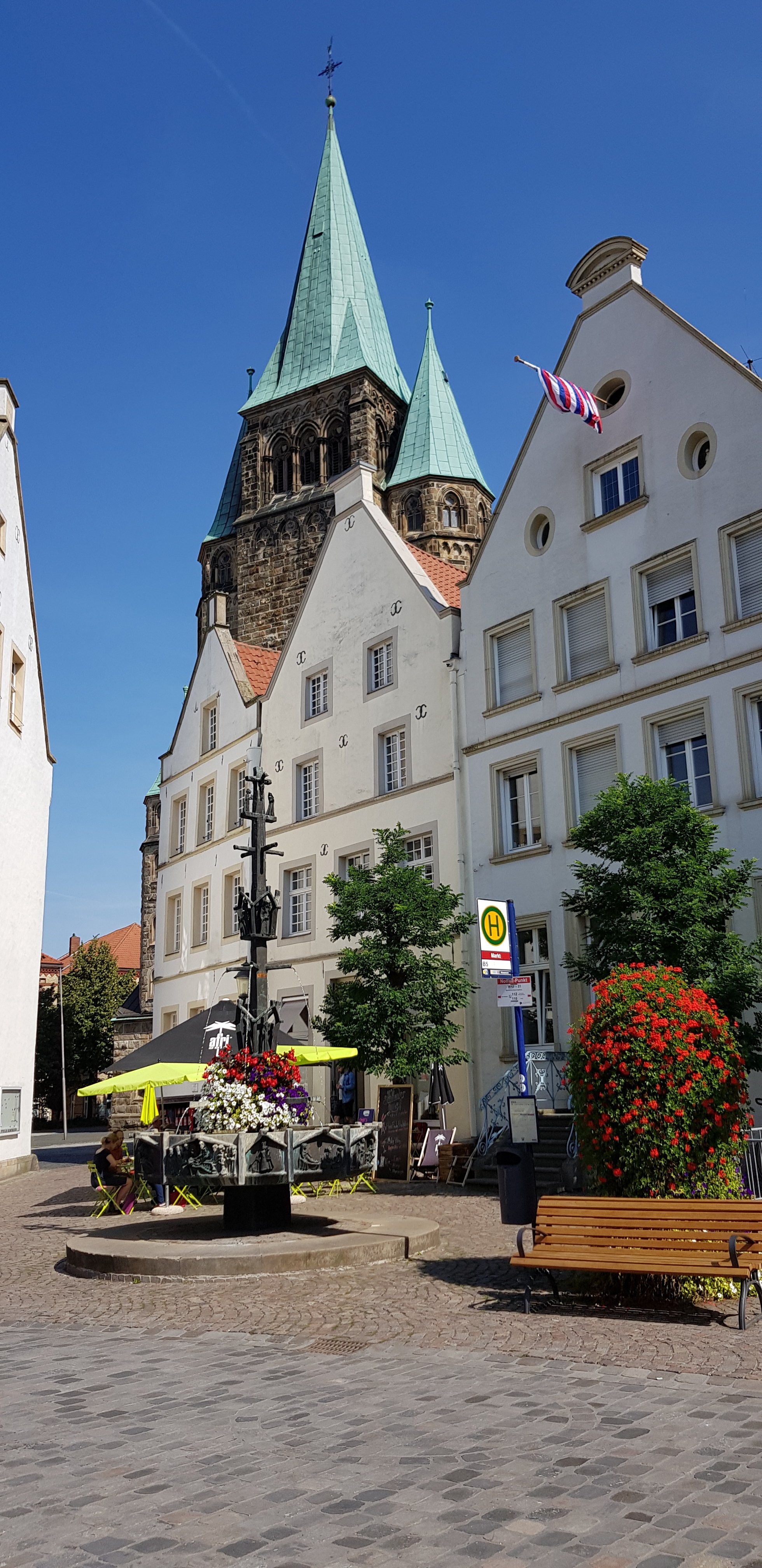 A sunny square featuring a decorative fountain with flowers, surrounded by buildings with white façades and a church tower in the background topped with a green spire.
