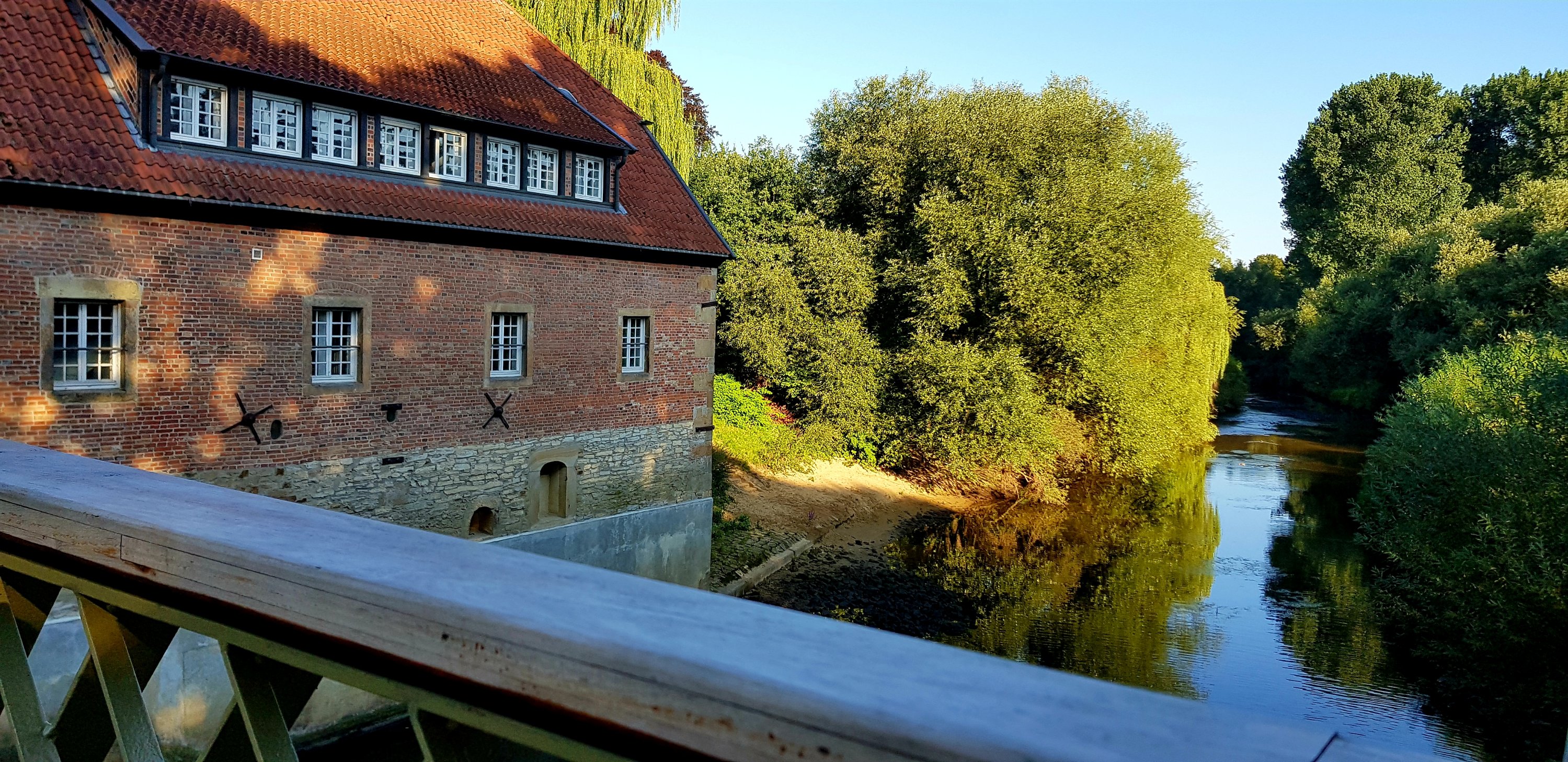 A scenic view of a brick building with a red roof beside a flowing river, surrounded by lush greenery under a clear blue sky.