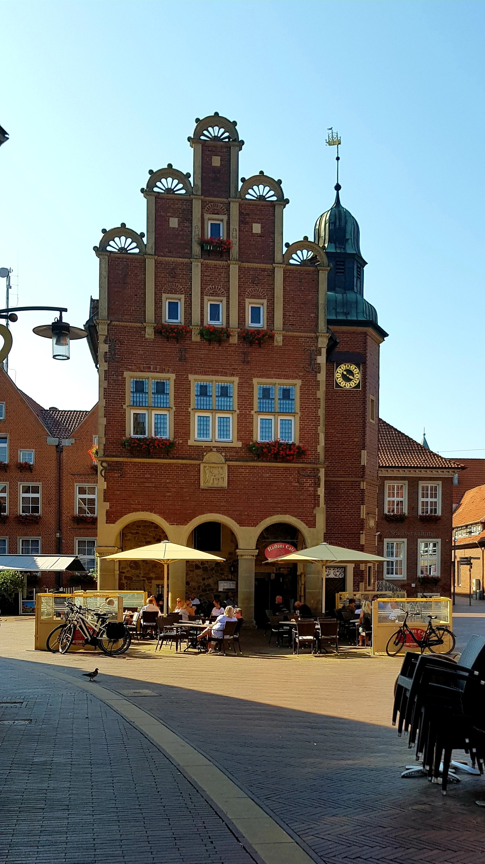 Historic building with intricate architecture, featuring a clock and flower boxes, situated in a sunny square with outdoor seating and bicycles.