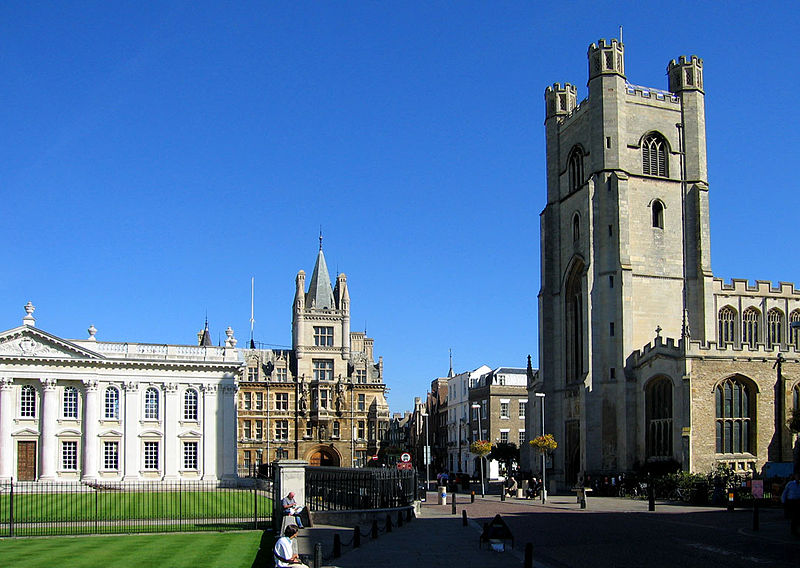 A panoramic view of historic buildings in Cambridge, featuring a white classical structure on the left and a tall stone church on the right, set against a clear blue sky.