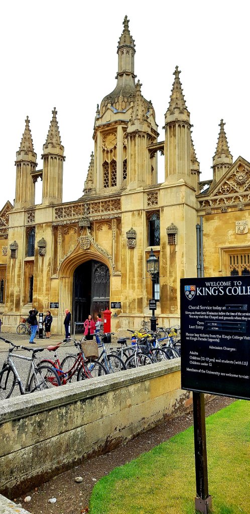 Facade of King's College with Gothic architecture, featuring spires and a central entrance. A sign welcomes visitors, while bicycles are parked nearby and people are gathered in front of the college.