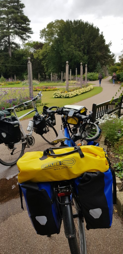 Bicycles parked in a garden with colorful flowers and walking paths in the background. The bike in focus features Ortlieb waterproof panniers.