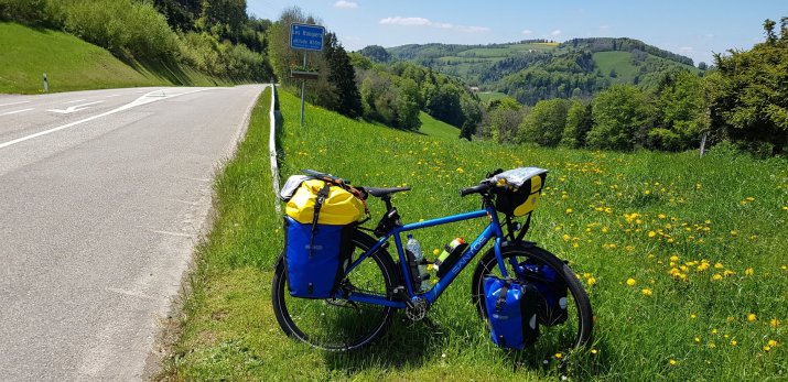A blue bicycle with yellow panniers is parked on the roadside near lush green grass and yellow dandelions, with a scenic view of rolling hills in the background.