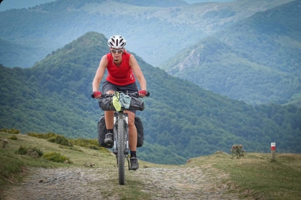 A cyclist riding a mountain bike on a gravel path surrounded by green hills and mountains.