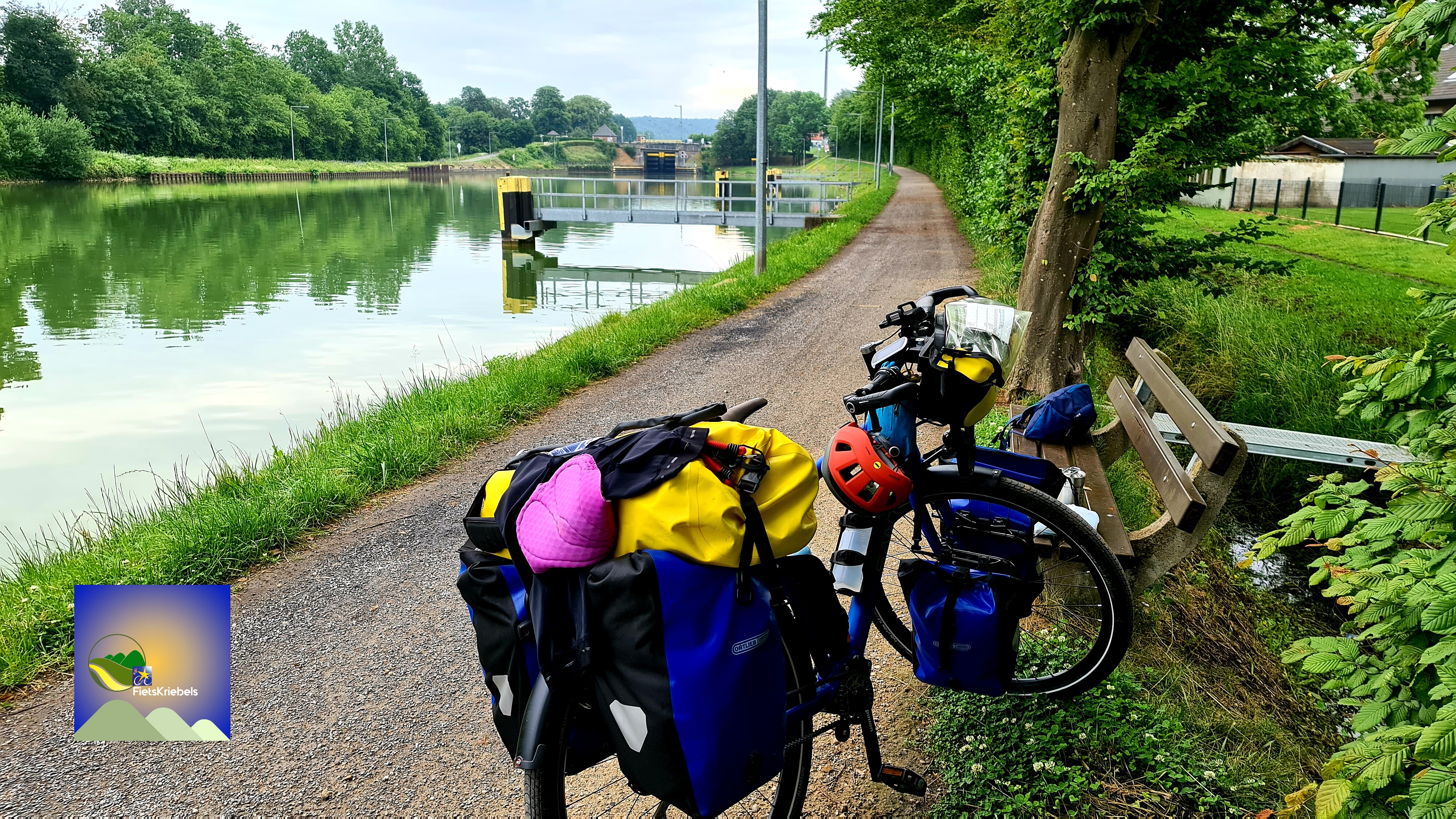A scenic view of a bike parked along a canal path, surrounded by lush greenery. The bike is equipped with colorful panniers and a helmet, while a bench is visible nearby, along with a reflection of trees in the calm water.