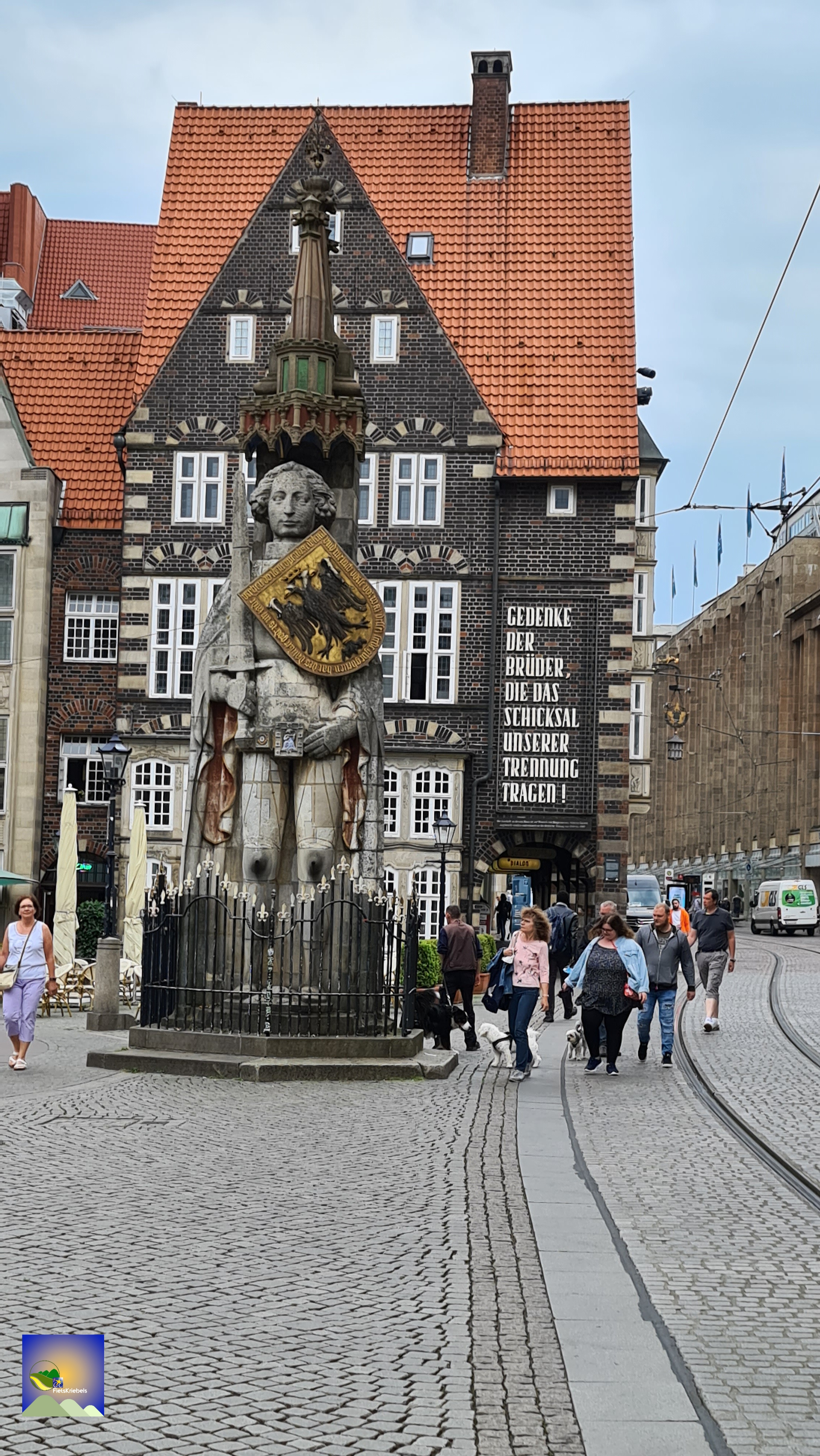 A pedestrian street with a large statue in the foreground featuring a figure holding a shield. Behind the statue is a historic building with black and white architecture and red tiled roofs. People walk by, with some taking dogs for a stroll.