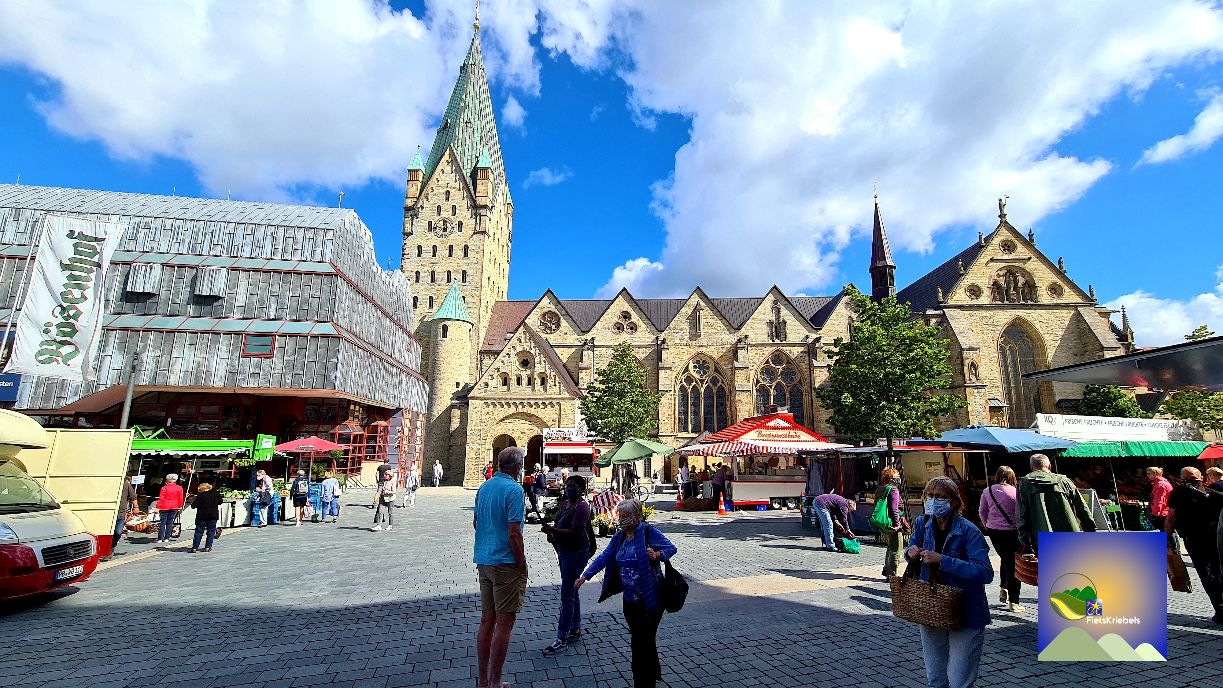 A bustling market square featuring historic buildings, including a tall stone church with a green-tipped tower. People are shopping at various stalls, with colorful awnings and food vendors.