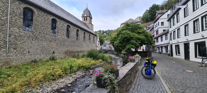 A scenic view of a cobblestone street leading to a historic stone building with large windows, alongside a small stream lined with greenery and flower pots. A bicycle with yellow and blue bags is parked near the water.