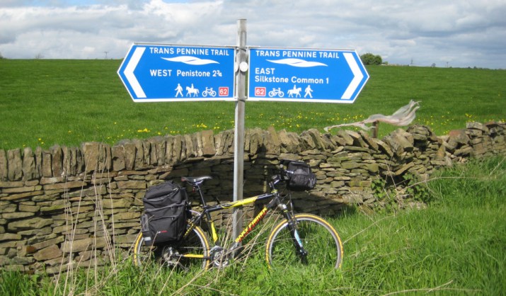 A bike leaning against a stone wall next to a signpost indicating directions for the Trans Pennine Trail, with destinations west to Penistone and east to Silkstone Common.