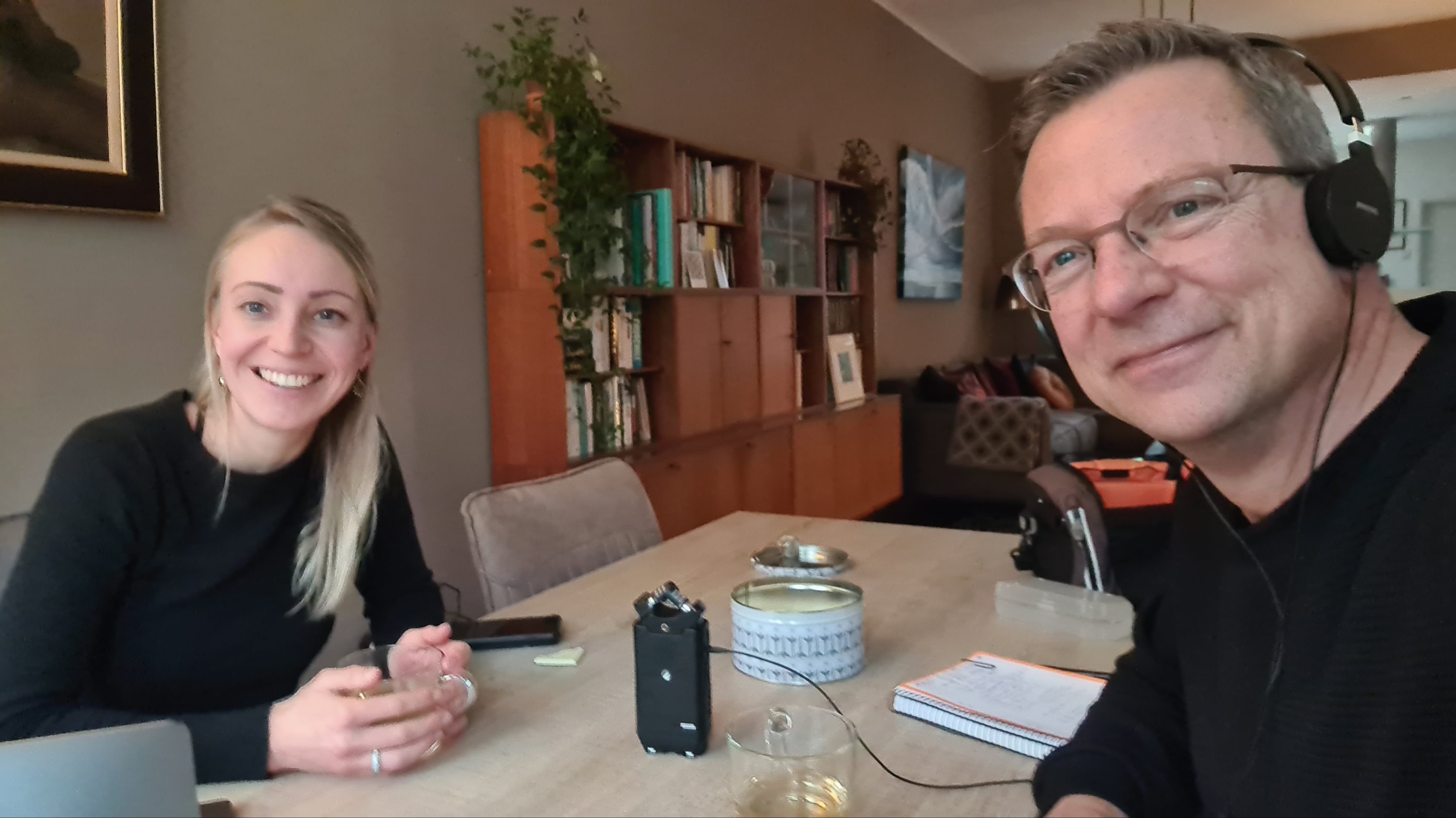 Twee mensen zitten aan een tafel in een huiselijke omgeving met boekenplanken op de achtergrond, één van hen houdt een glas vast en beiden glimlachen naar de camera.