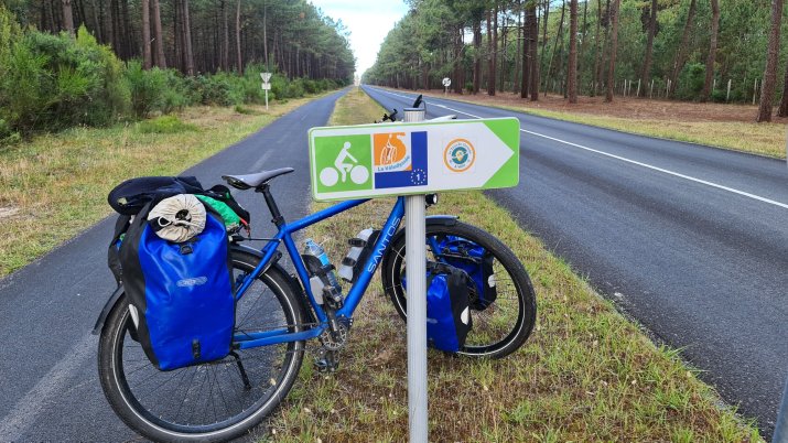 A blue bicycle with saddlebags parked beside a directional sign for cyclists on a tree-lined road. France Atlantique - West Frankrijk Route