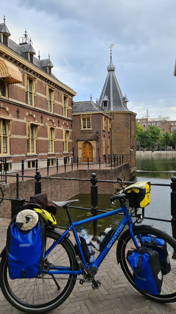 A blue bicycle with bags parked near a canal, with a historical brick building and a tower in the background under a cloudy sky.