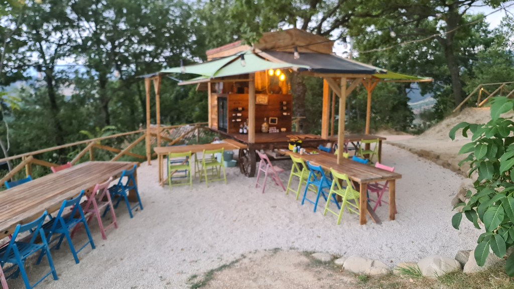 Een houten eetwagen met overdekte zitplaatsen en kleurrijke stoelen, omgeven door groen, op een gravelpad.