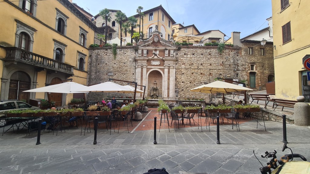 A charming Italian square featuring an old stone fountain, surrounded by tables with umbrellas and greenery, with historic buildings in the background.