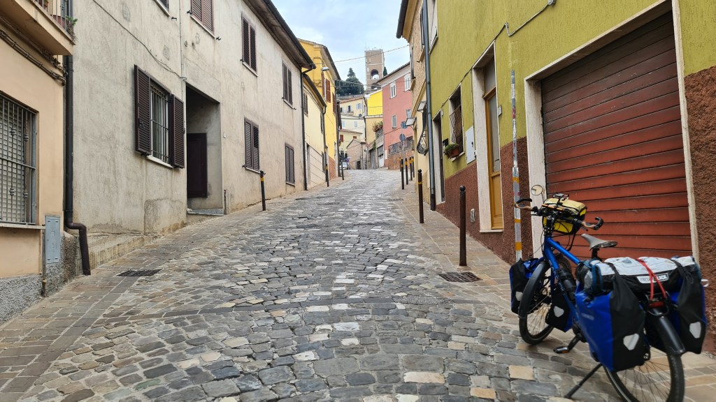 A cobblestone street winding uphill flanked by colorful buildings and a parked bicycle with blue and black panniers.