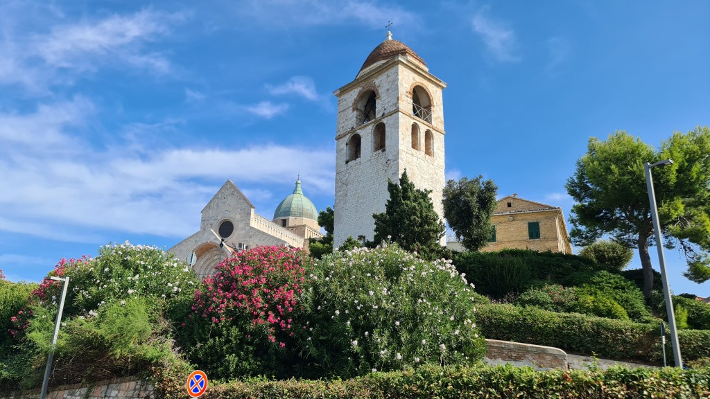 Historic church tower surrounded by lush greenery and colorful flowers against a blue sky.
