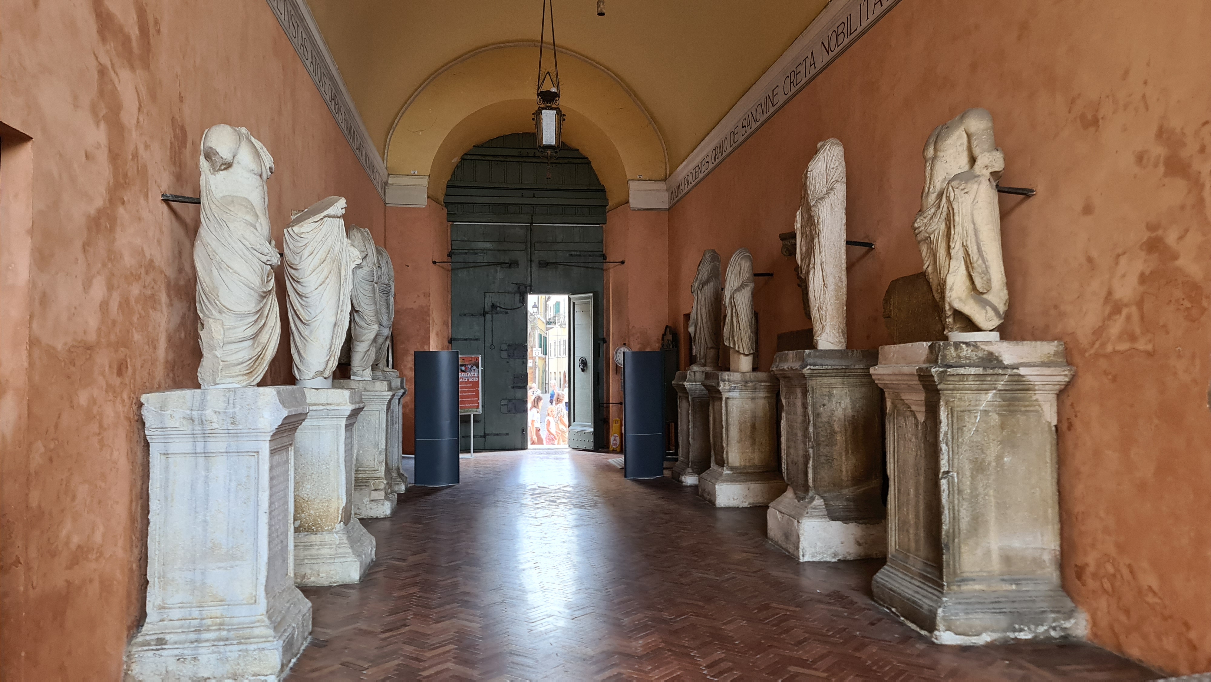 Een gang met marmeren beelden en een open deur aan het einde, met een warme, oranje achtergrond.